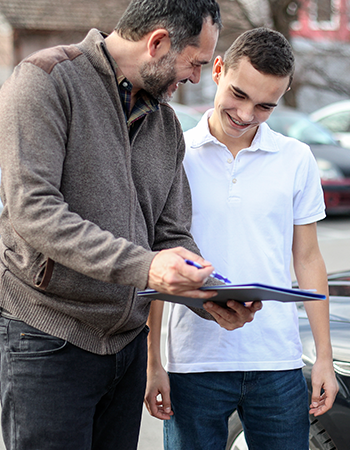 A driving instructor looking at a clipboard alongside a male teen driver