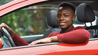 A teen boy in a red sweater sits in the driver seat of a red car and smiles at the camera.