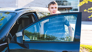 A teen boy carries a backpack as he gets in the drivers seat of a car parked near a school