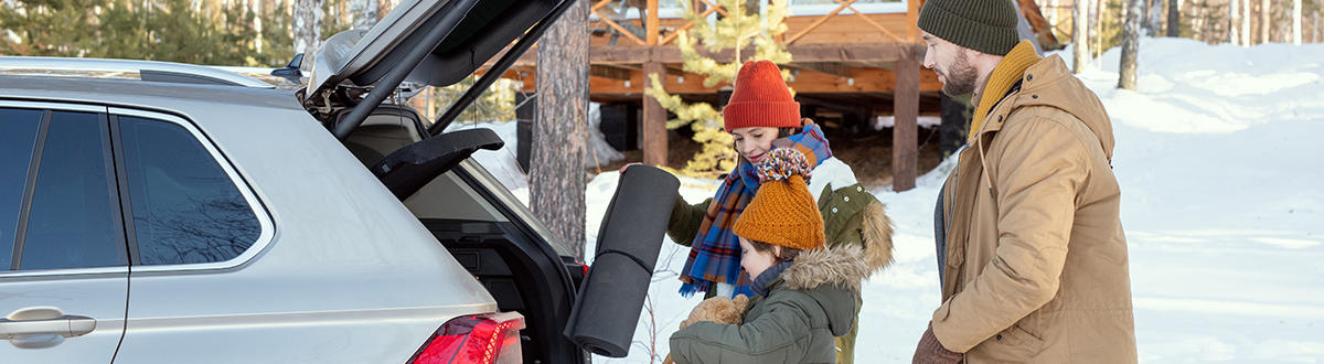 Family of three in warm winter clothing loading luggage into the back of a vehicle in snowy weather