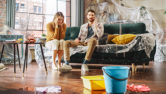 A sad couple sit on a plastic-covered couch as water drips from the ceiling due to a leak upstairs.