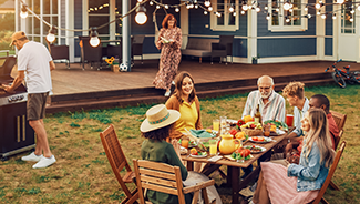 Family and friends are celebrating with an outdoor gathering in the yard of a large house. A man grills, a group sits at a table, and children play.