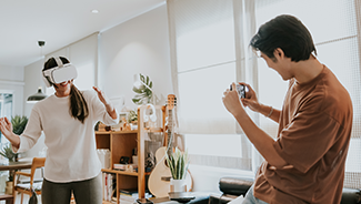 A woman in a cream sweater is playfully modeling a new VR headset as a man, in jeans and an orange t-shirt, photographs the item for their home inventory record.