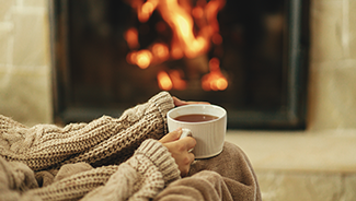 Hands in a cozy sweater hold a cup of warm tea with a burning fireplace in the background