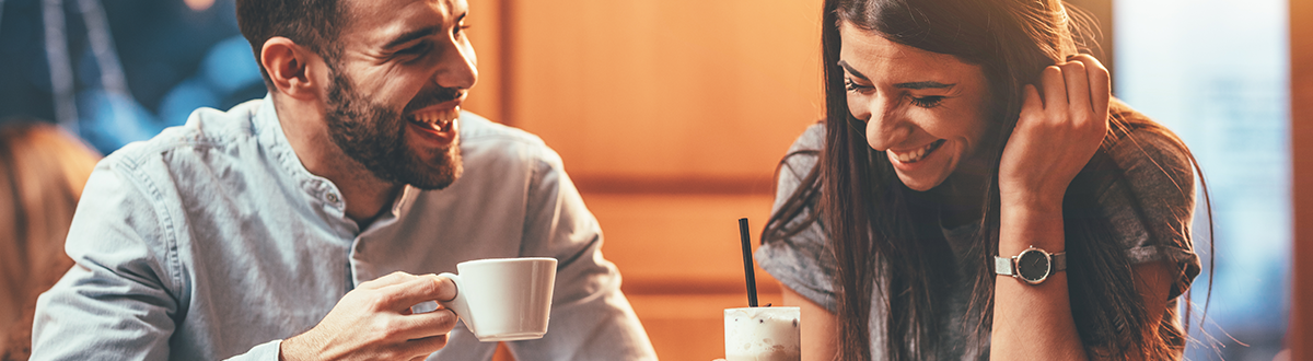 A man, wearing a white denim shirt, and a woman, wearing a gray T-shirt, are drinking coffee together at a cafe.