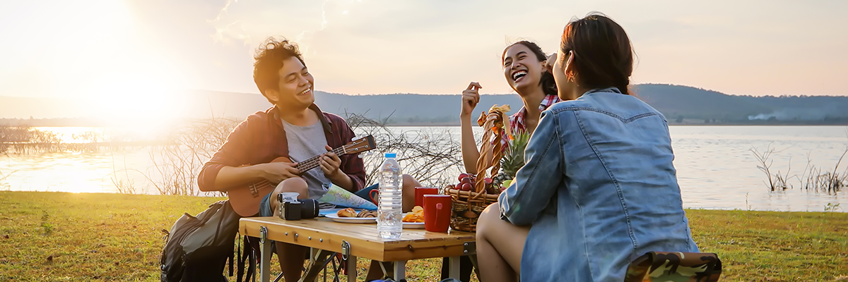 Three friends (one playing a guitar) have fun at a summer picnic near a lake at sunset.