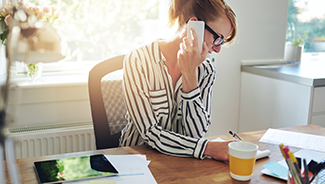 A woman at a desk holds a phone to her ear in one hand while she writes on a notepad with the other.