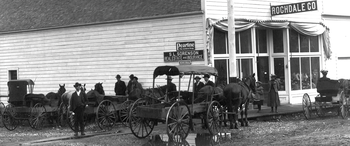 Black and white photo of Enumclaw, WA in the 1800's. Along with horse drawn carriages, the image features a sign for S.L. Sorenson Real Estate and Insurance. Sorenson was the first agent at what would later become Mutual of Enumclaw Insurance.