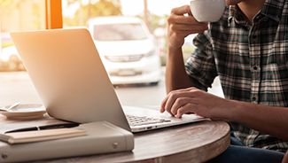 A man in a flannel shirt is sipping coffee while working on a laptop.