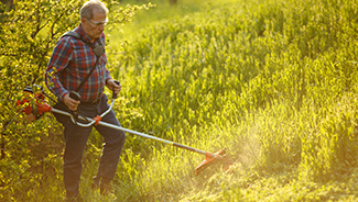 A man using a hand-held brush trimmer to shorten tall grass around the perimeter of his property